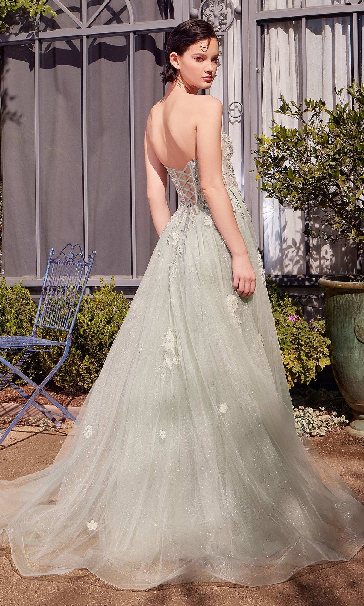 A woman wears the Andrea & Leo A1418 sage green prom ball gown with floral embellishments. The strapless corset dress has a lace-up back and sweeping train as she stands outdoors, glancing over her shoulder.